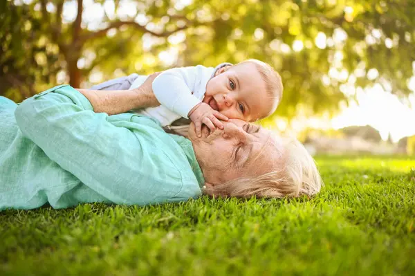 Beautiful happy smiling senior elderly woman lying on grass with cute little baby boy. Grandmother and grandson having fun time together at tropical summer day park. Multigenerational family concept
