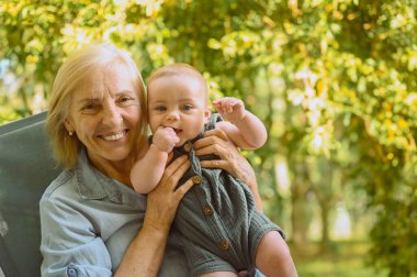 Güzel, mutlu, gülümseyen yaşlı bir kadın. Ellerini tutuyor. Dışarıda oturan sevimli bir erkek bebek. Büyükannenin torunu güneşli bir yaz gününde bahçede ya da parkta eğleniyor.