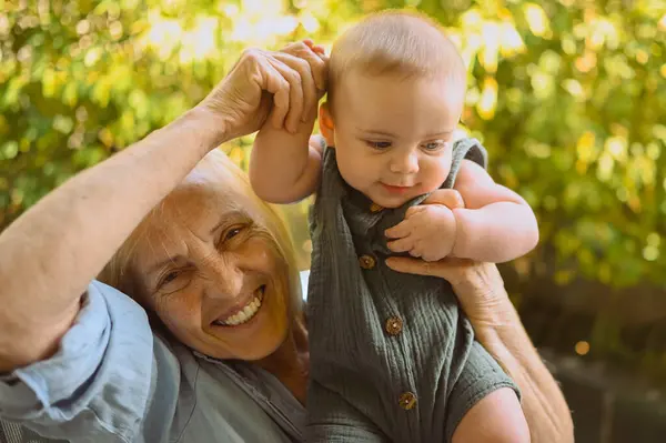 Beautiful happy smiling senior elderly woman holding on hands cute little baby boy sitting outdoor. Grandmother grandson having fun time together at sunny summer day at garden or park