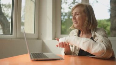 Yaşlı bir kadın diz üstü bilgisayarla görüntülü sohbet yapıyor. Kırık kolu içeride, pencerelerin önünde. Alçıtaşlı bandajla bileğe dokunan yaşlı kadın acı çekiyor.. 