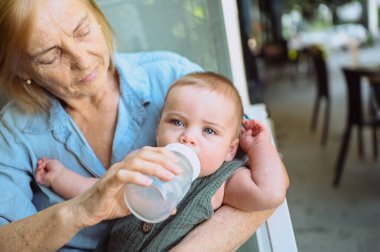 Güzel, mutlu, gülümseyen, yaşlı, yaşlı bir kadın dadının mama formülünü küçük, tatlı bir erkek bebekten yedirmesi. Büyükannem ve torunum yaz günü kaldırımdaki kafede. Çok nesilli aile kavramı