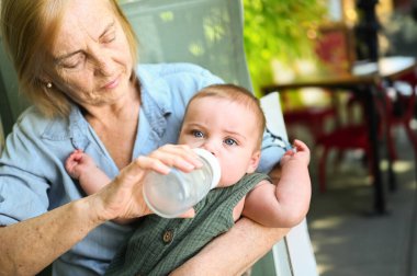 Güzel, mutlu, gülümseyen, yaşlı, yaşlı bir kadın dadının mama formülünü küçük, tatlı bir erkek bebekten yedirmesi. Büyükannem ve torunum yaz günü kaldırımdaki kafede. Çok nesilli aile kavramı