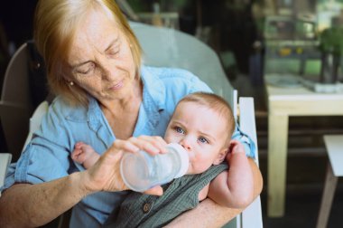 Güzel, mutlu, gülümseyen, yaşlı, yaşlı bir kadın dadının mama formülünü küçük, tatlı bir erkek bebekten yedirmesi. Büyükannem ve torunum yaz günü kaldırımdaki kafede. Çok nesilli aile kavramı