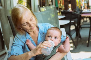 Güzel, mutlu, gülümseyen, yaşlı, yaşlı bir kadın dadının mama formülünü küçük, tatlı bir erkek bebekten yedirmesi. Büyükannem ve torunum yaz günü kaldırımdaki kafede. Çok nesilli aile kavramı