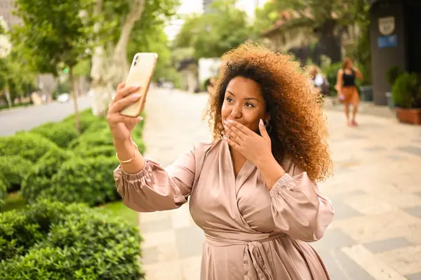 Attractive happy stylish plus size African American woman student freelancer afro hair making selfie with smartphone outside. Young lady in silk dress in big urban city street at summer. Diversity.