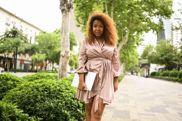 Attractive happy stylish plus size African American woman student freelancer afro hair with laptop computer outside, big urban city street at summer. Diversity. Remote office work, distance education