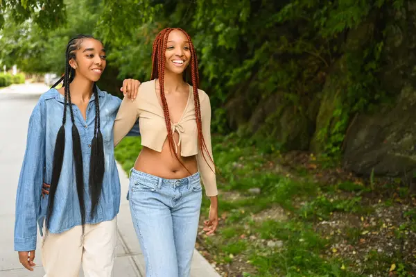 Sokak tarzı güzel Afro-Amerikan kadın siyah çift portresi. Saç örgüsü, mükemmel beyaz dişler, kız kardeş arkadaşlar güneşli bir yaz gününde dışarıda yürüyorlar. Yeşil park ve şehir