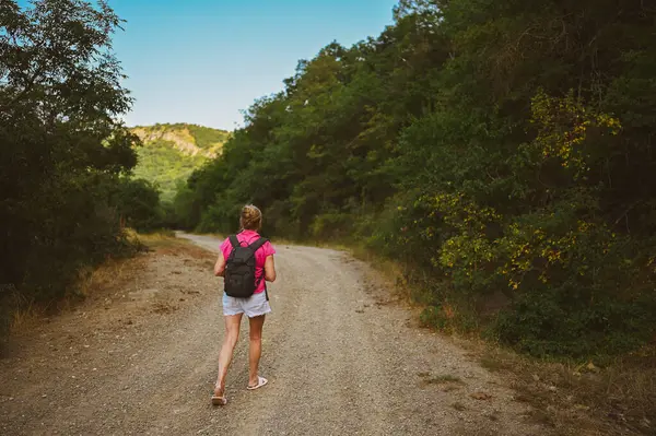 Kıdemli kadın sırt çantalı turist yaz ormanı yolunda gün batımında arka planda yürüyor. Yaşlı sıska bayan yalnız seyahat ediyor. Aktif emeklilik tatili ve sağlıklı yaşam tarzı konsepti. Sıcak filtre.