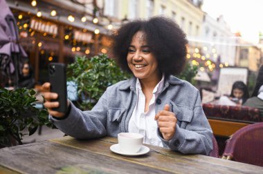 Street style portrait of attractive young happy African American woman with afro hair have video chat with smartphone outdoors in sidewalk cafe. Excited smiling female blogger broadcasts live stream