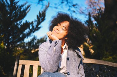 Close up Fashion street style portrait of attractive young natural beauty African American woman with afro hair in blue coat posing walking outdoors in sunny day. Happy lady with perfect teeth smile