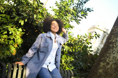Fashion street style portrait of attractive young natural beauty African American woman with afro hair in blue coat and jeans posing outdoors. Happy tourist laughing walks around the city