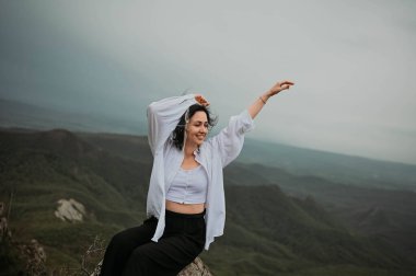 Mutlu kadın gülümseyen turist dağda yürüyüş yapıyor ve doğanın tadını çıkarıyor. Bulutlu, kasvetli, rüzgarlı bir havada manzara. Beyaz gömlekli, kolları açık, zevk içinde gezmekten hoşlanan bir kadın. Özgürlük kavramı