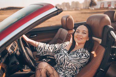 Kumsal kıyısındaki yeni kırmızı bir arabada oturan, gülümseyen esmer kadın sürücü kameraya bakıp yolculuğun tadını çıkarıyor. Sürücü kursları ve hayat sigortası konsepti. Göz kamaştırıcı yaşam tarzı