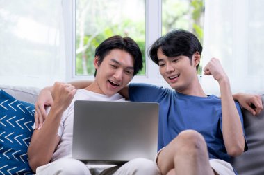 Oturma odasında dizüstü bilgisayar kullanan mutlu Asyalı eşcinsel çift. Mutlu bir şekilde ellerini kaldırarak zaferi, başarıyı ya da iyi haberi kutluyorlar.