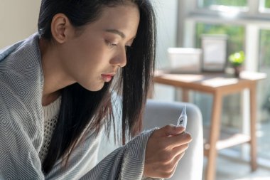 Evdeki kanepede yatan hasta genç Asyalı kadın battaniyeyle örtüyor ve termometreyle ateşini ölçüyor.