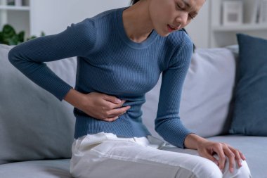 Genç Asyalı kadın evdeki kanepede otururken karın ağrısı çekiyor. Hasta kadının midesi ağrıyor..