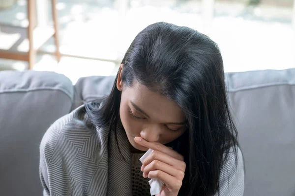 Ill young Asian woman covered with blanket blowing running nose got fever caught cold sneezing in tissue sit on sofa, sick allergic girl having allergy symptoms coughing at home