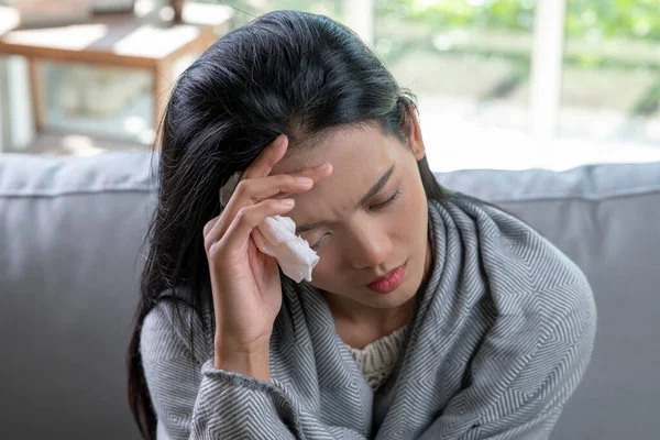 Young Asian woman covered with blanket having headache from flu illness sit on sofa, sick allergic girl having allergy symptoms coughing at home