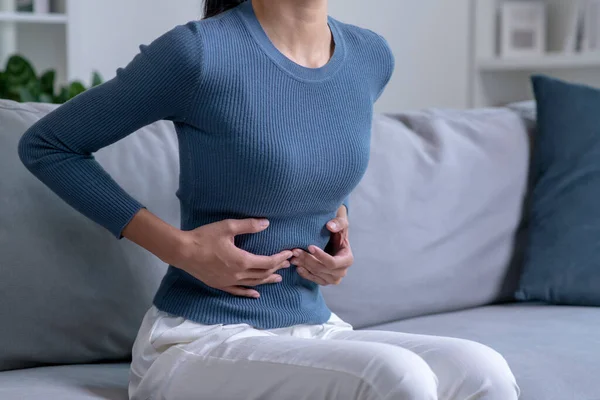 stock image Young woman suffering from strong abdominal pain while sitting on sofa at home. Sick female having a stomach ache.