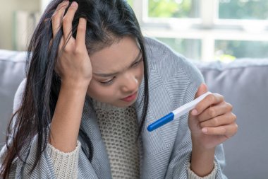 Depresyondaki genç Asyalı kadın elinde negatif ya da pozitif hamilelik testi tutuyor..