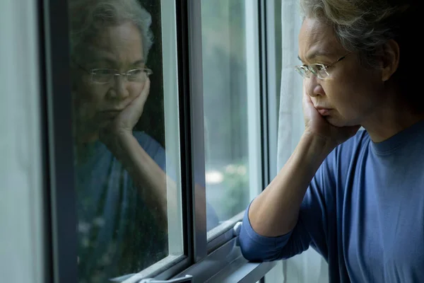 一位忧心忡忡的亚洲老年妇女从窗户往外看的画像 — 图库照片