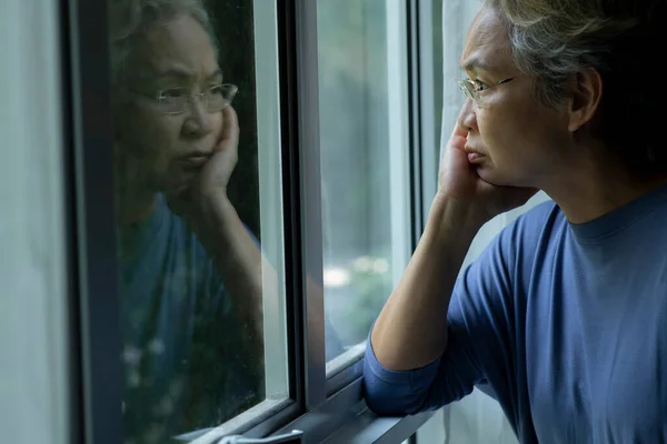 Portrait of sad and worried Asian senior woman looking through the window at home