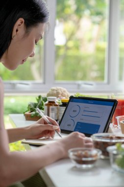 Masada çeşitli meyveler, fındıklar, sebzeler ve beslenme malzemeleri bulunan bir dizüstü bilgisayardan veri toplayan Asyalı kadın beslenme uzmanı.