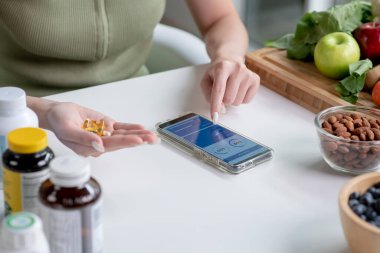 Kadın profesyonel beslenme uzmanı elinde besin takviyelerini kontrol ediyor. Masada çeşitli meyveler, fındıklar, sebzeler ve beslenme malzemeleri var.