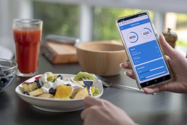 Yemek yerken telefonundaki kalori ve besin uygulamalarını kontrol eden bir kadın. Profesyonel Beslenme Uzmanı.