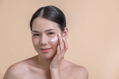 Güzel Asyalı kadın yüzüne nemlendirici krem sürüyor. Bej arka planda mükemmel makyajlı gülümseyen bir kadının fotoğrafı. Güzellik konsepti