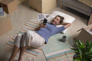 Genç Asyalı bir kadının taşındıktan sonra yeni evinde yerde yatıp akıllı telefon kullanmasının en iyi görüntüsü..