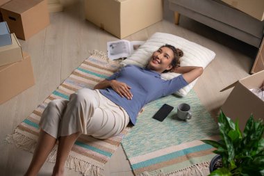 Yeni evde dinlenen mutlu Asyalı genç bir kadın. Taşındıktan sonra gülümseyen yüzüyle yerde yatıyor..