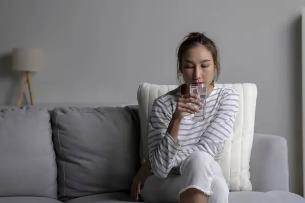 Asian woman drinking cold water while sitting on the sofa at home. Female drinking clean water health.