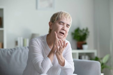Diş ağrısı olan Asyalı olgun bir kadın. Yaşlı yaşlı kadının dişi ağrıyor. Evdeki kanepede oturmuş, diş ağrısı çeken mutsuz bir kadın kendini kötü hissediyor. Çenesini tutan üzgün, yaşlı bir kadın. Yetişkinler diş ağrısı çekiyor.