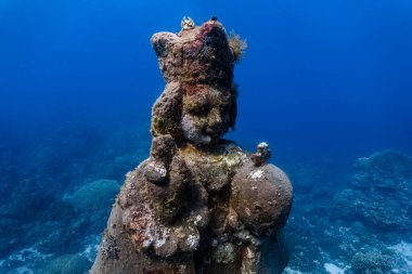 Okyanus tabanındaki bir yerde deniz canlılarıyla çevrili sakin bir figürün sualtı heykeli.