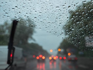 Closeup of raindrops on a windshield, blurry wipers cleaning the glass, and road traffic with blurry car lights. View from the driver's seat.