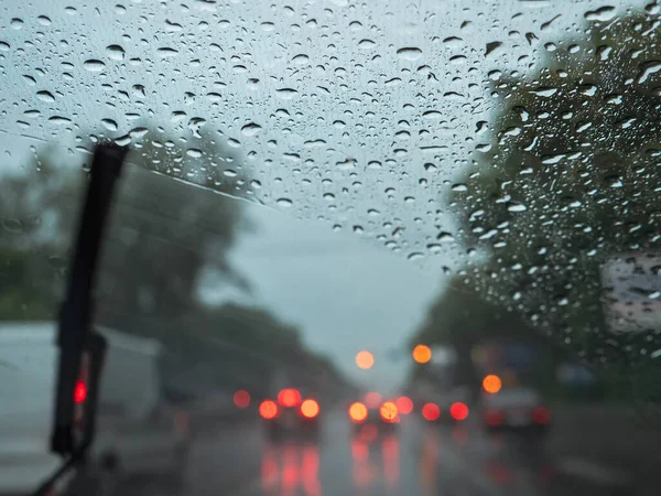 Closeup of raindrops on a windshield, blurry wipers cleaning the glass, and road traffic with blurry car lights. View from the driver's seat.