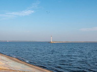Kyiv denizinin kıyısındaki eski beton deniz feneri üzerinde uçan kuşlar, beyaz yatlı sakin mavi su ve ufuktaki dar banka şeridi ile mavi gökyüzü..