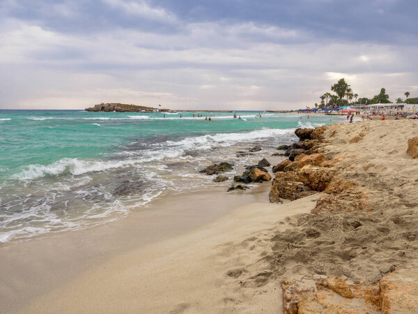Люди плавают в морских волнах в пасмурный день. Живописный пляж Нисси с мутной голубой водой, маленьким скальным островом и темным облачным небом в Айя Фаста, Кипрус. Восхитительные морские каникулы
.