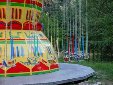 Şehir parkında eski usul bir atlıkarınca. Sallanan atlıkarıncanın metal zincirlerinde kırmızı ve mavi tahta koltuklar. Arka planda ağaçlar, yerde düşen yapraklar..