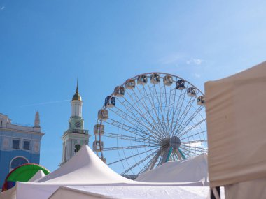 Modern observation wheel and ancient church bell tower with bright blue cloudless sky in the background in Kontraktova square, Podil, Kyiv city. Travelling and entertainment in old city center. clipart