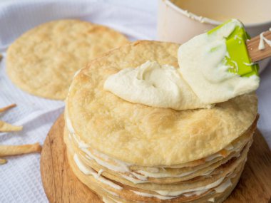 Silicone spatula spreading vanilla custard over a pile of baked thin round buttery layers on a wooden board, a bowl and a new pile waiting in the background ready to make a shortcrust puff pastry Napoleon cake. clipart