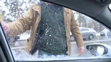 Uzun saplı kar fırçalı yeşil kapüşonlu bir adam ön cam ve yan camlardan buz ve kar kaldırıyor. Sürücü koltuğundan görüntü.