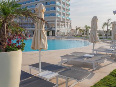 Güneşli yataklardan, kapalı şemsiyelerden ve yüzme havuzunun üzerinde bir palmiye ağacından ve açık hava restoranı olan ve Kıbrıs Rum Kesimi 'nin Ayia Napa kentinde kahvaltı yapan insanlarla dolu bir otel binasından manzara.