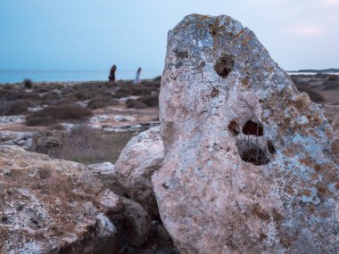 Pençe izi gibi büyük delikleri olan dev gözenekli bir kayanın yakınındaki Akdeniz kıyısında dikey duruyor, kayalık kıyılarda ve Kıbrıs Rum Kesimi 'nin Ayia Napa şehrinde arka planda yürüyen iki kadın.