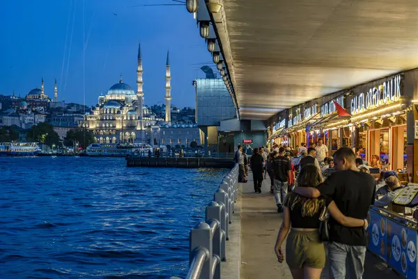 Hindi. İstanbul. Galata Köprüsü 'ndeki gece sahnesinde sağda hareketli restoranlar ve arka planda aydınlık Yeni Cami var.