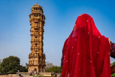 India. Rajasthan. Chittorgarh. Chittor Fort. An Indian woman in a red sari admires the Vijaya Stambha (Victory Tower) clipart