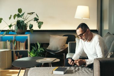 Evdeki kanepede akıllı telefon ve gözlüklü orta yaşlı bir adam..