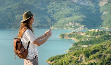 Şapkalı ve sırt çantalı gezgin Dağların ve gölün güzel manzarasının tadını çıkarıyor 
