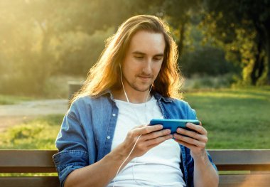 Genç adam parkta otururken cep telefonundan video izliyor.